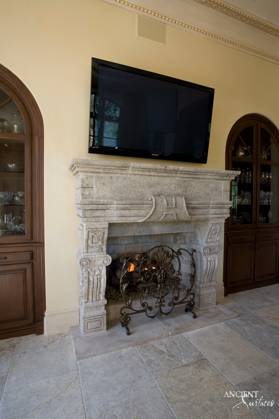 Reclaimed French antique limestone fireplace mantel from Ancient Surfaces with a carved centre cartouche and acanthus corbels installed beside arched cabinetry