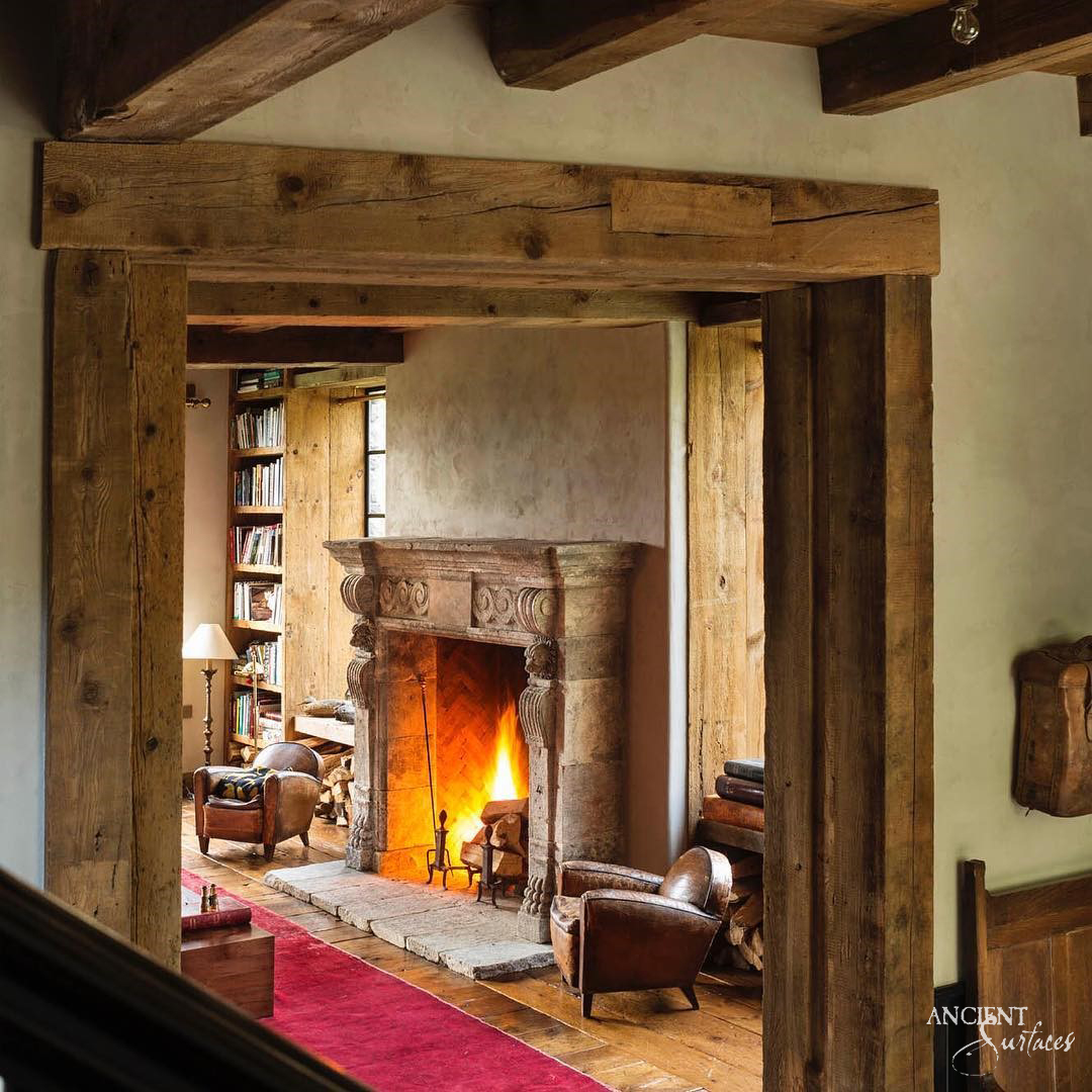 Ancient Surfaces reclaimed antique limestone fireplace mantel with carved column jambs and a herringbone firebox in a timber framed library