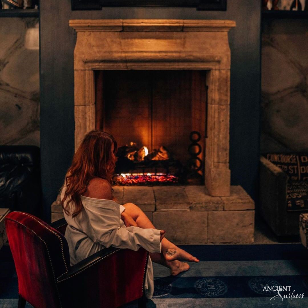 Reclaimed French limestone fireplace mantel from Ancient Surfaces on a raised stone hearth, lit by firelight in a dark panelled room